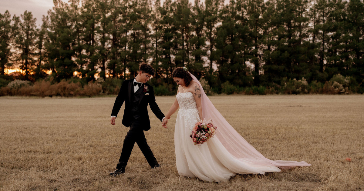 A man in a tuxedo and a woman in a white wedding dress and pink veil stand in front of pine trees at sunset.