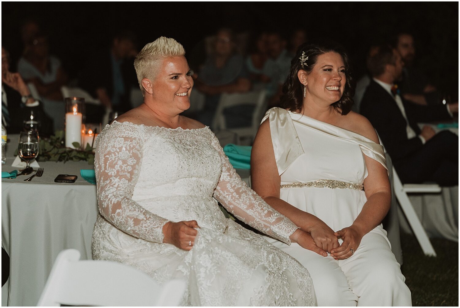 Two brides hold hands and smile during a speech.