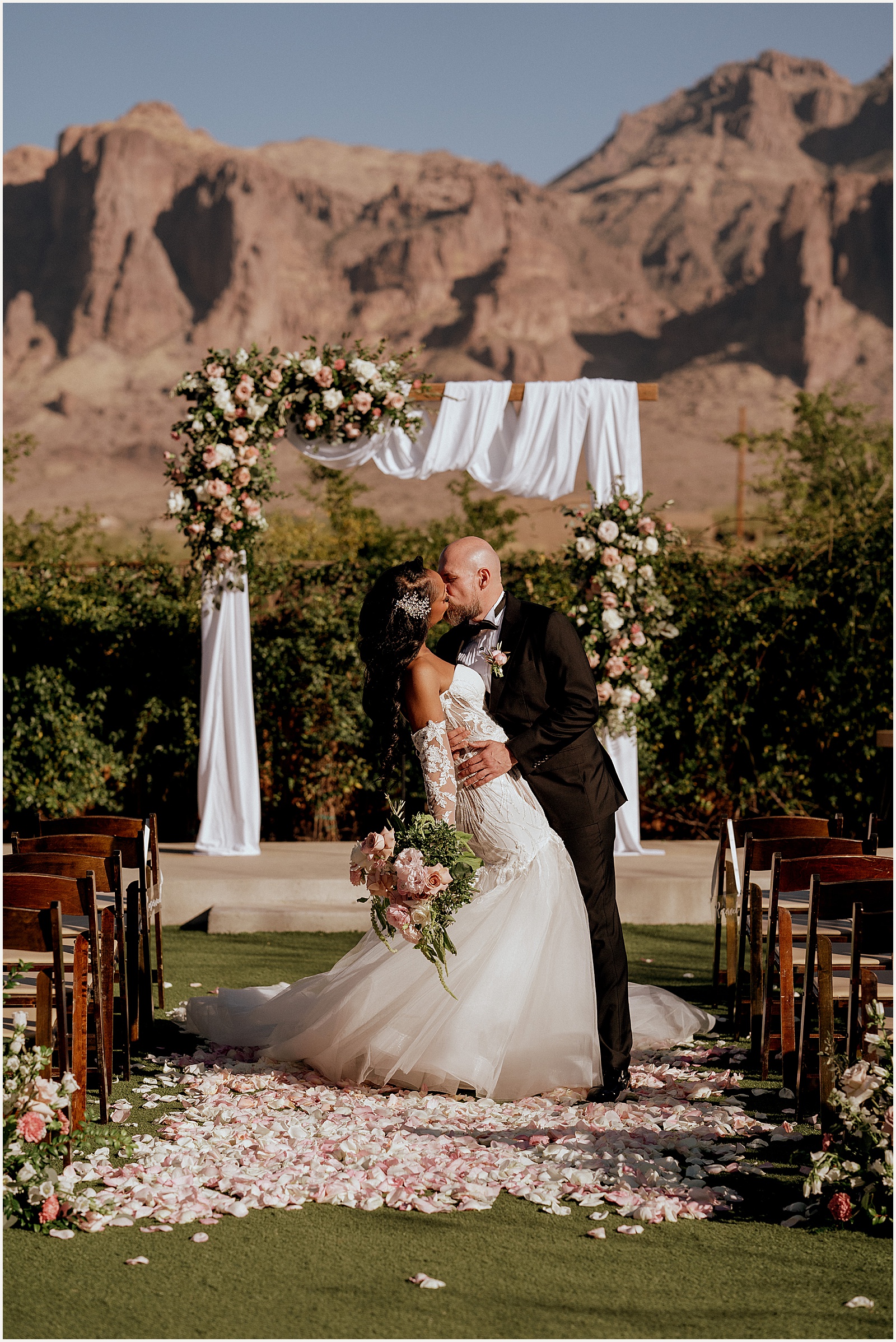 A bride and groom kiss in front of a floral arch at an Apache Junction wedding venue.