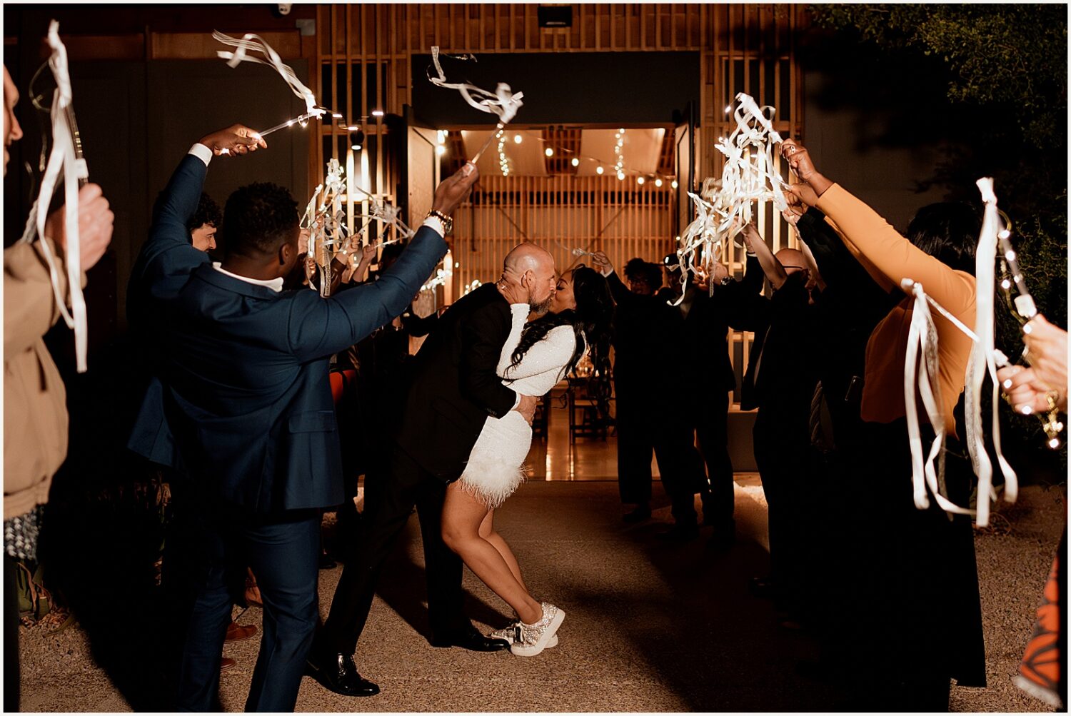 A bride and groom kiss as wedding guests wave sparklers at their wedding exit.