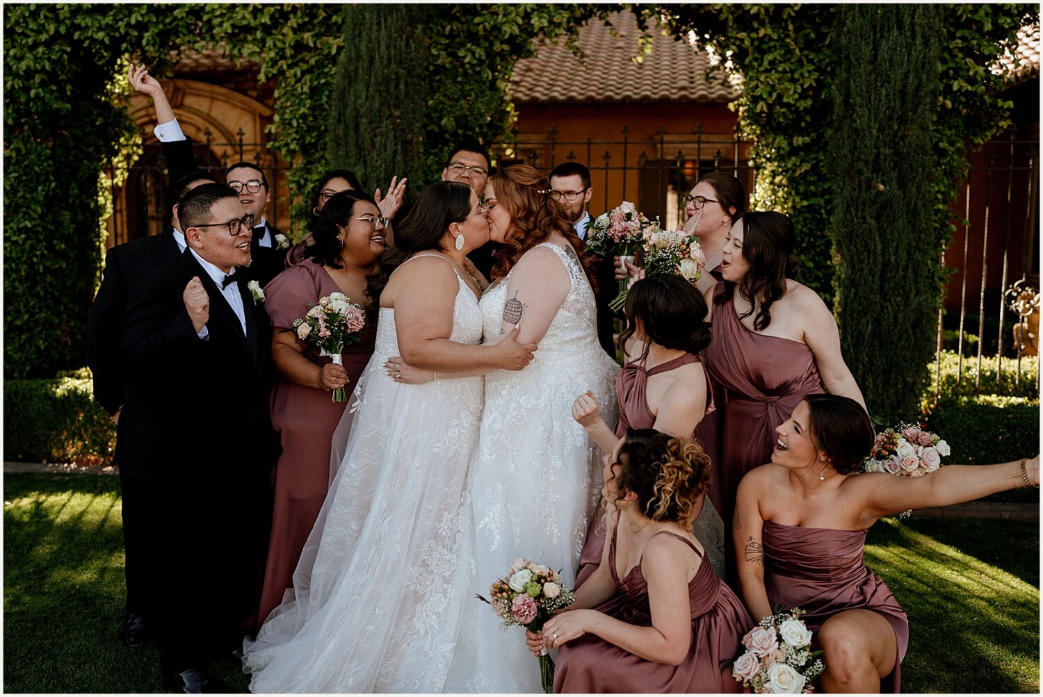 Two brides kiss surrounded by cheering bridesmaids.