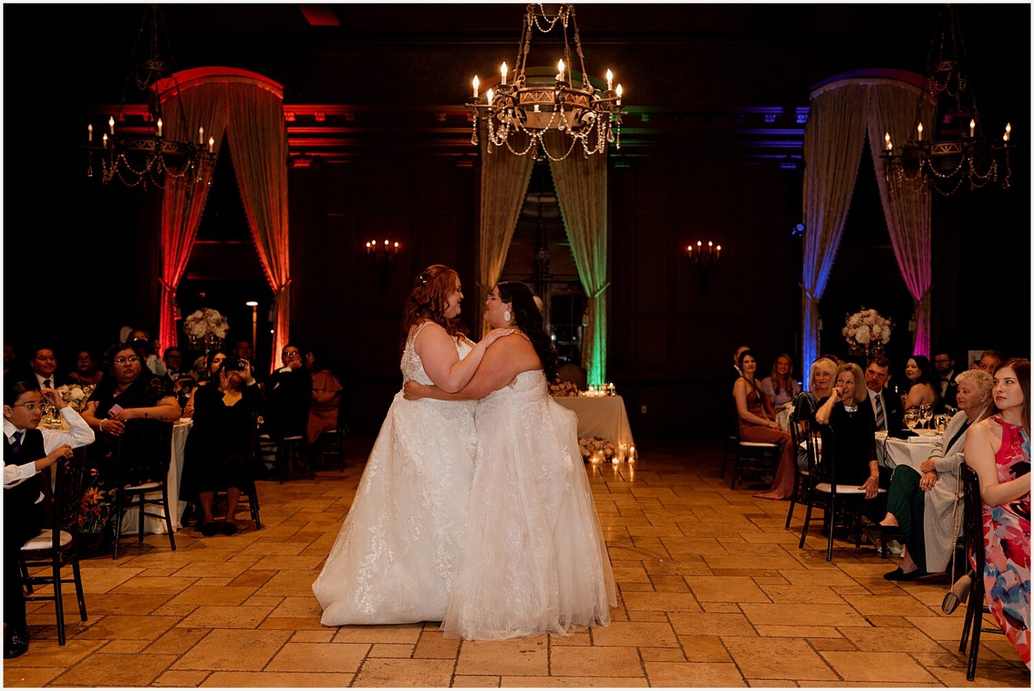 Two brides dance in a ballroom with rainbow uplighting.