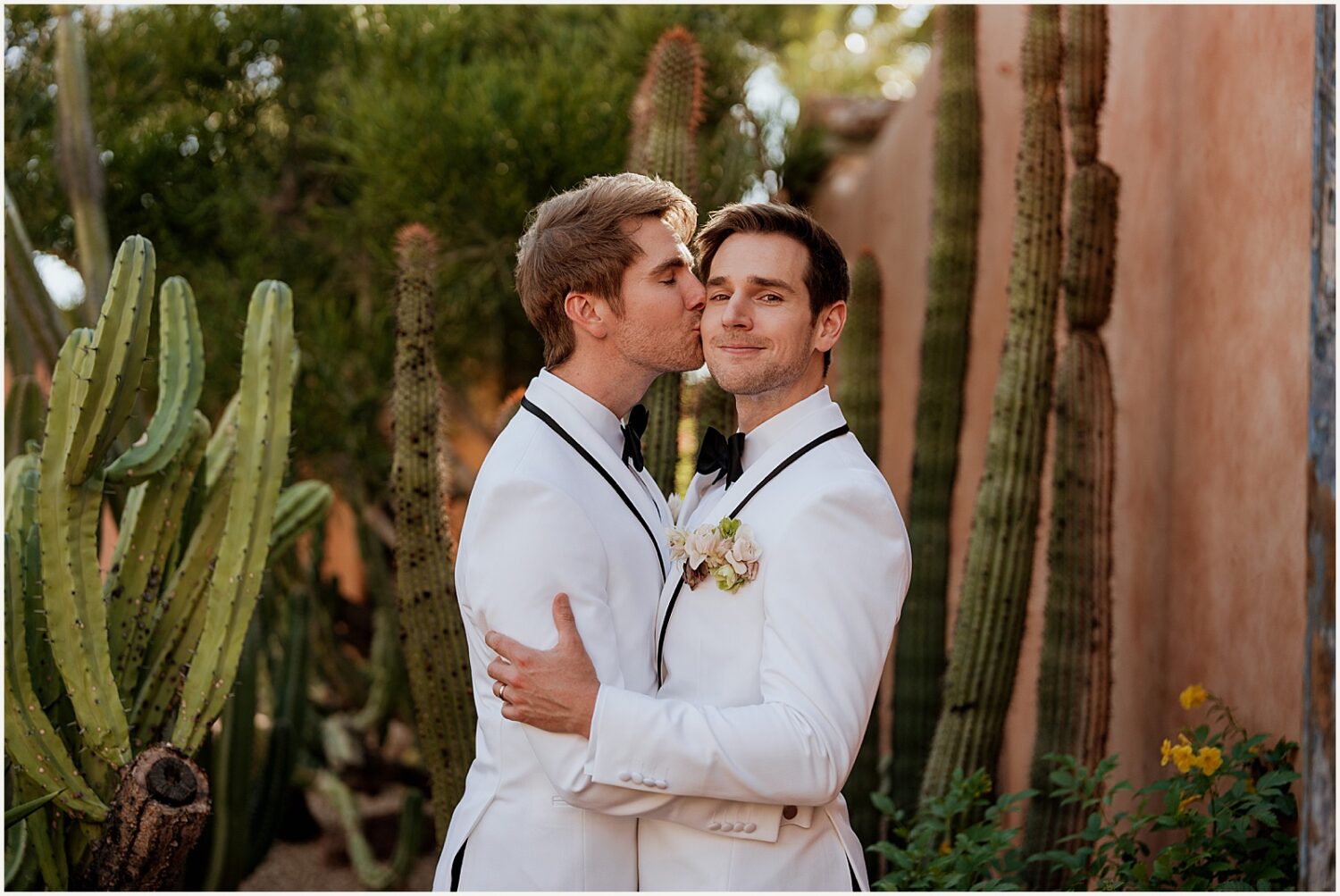 One groom kisses another on the cheek in a garden at an Arizona wedding venue.