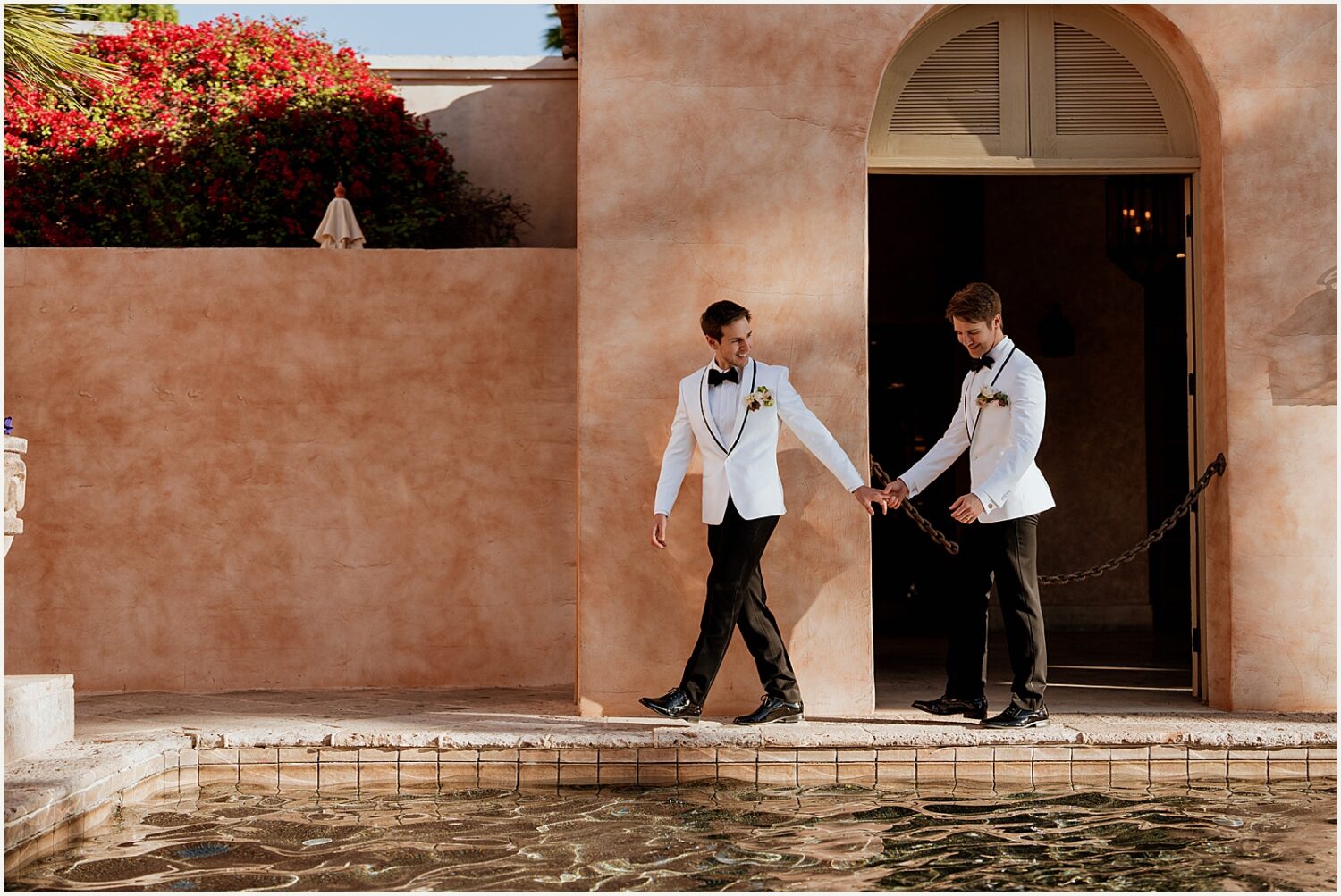 Two grooms walk down a poolside pathway holding hands.