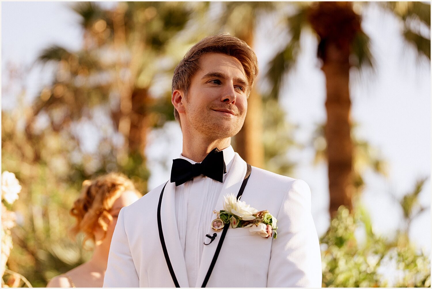 A groom stands under a palm tree smiling.