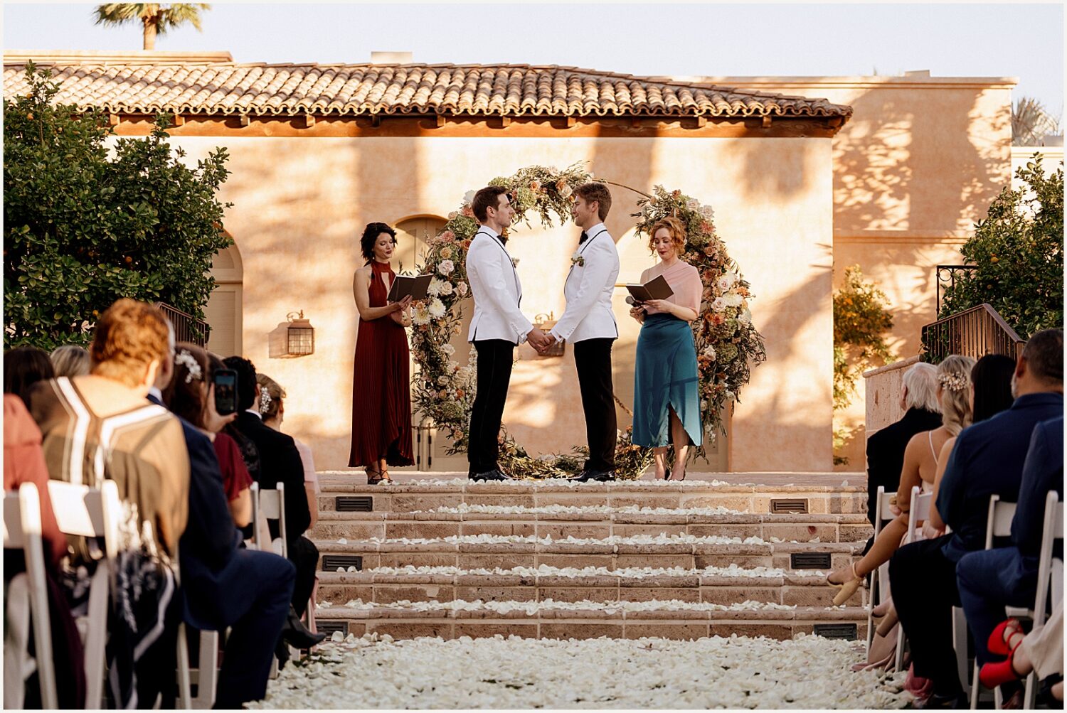 Two grooms stand in front of a floral arch at a Royal Palms wedding.