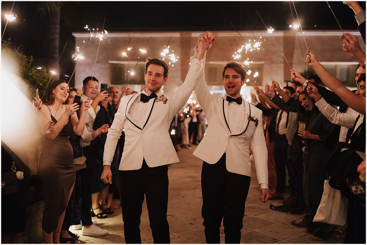 Two grooms raise their arms during a sparkler exit at an Arizona wedding.