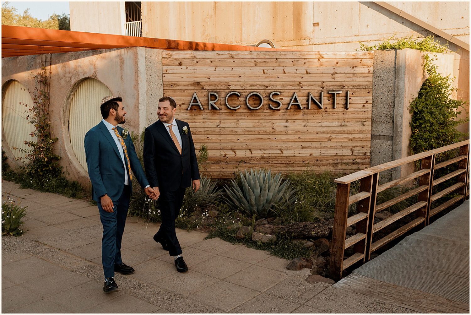 Two grooms hold hands beside the Arcosanti sign.