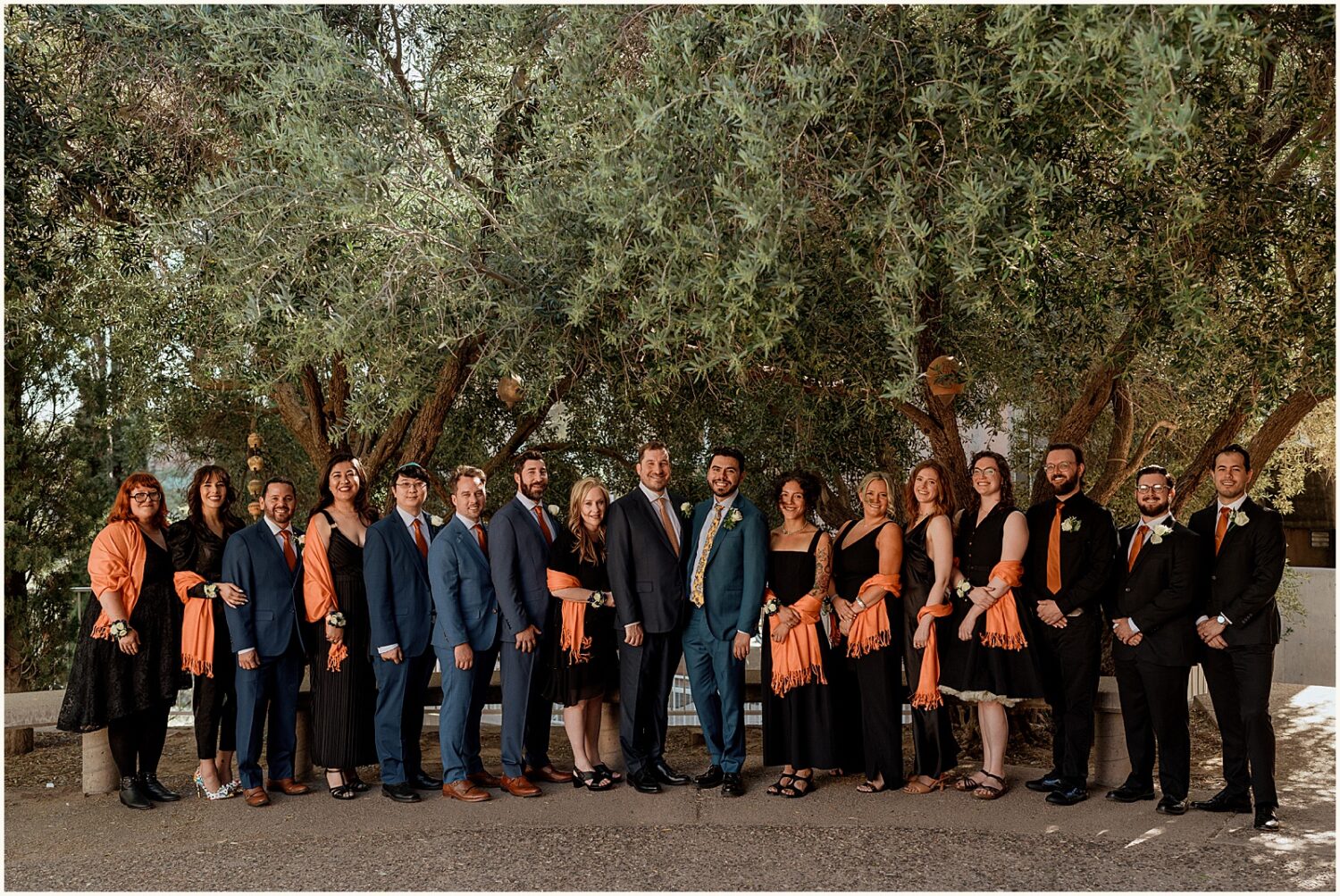 Two grooms pose with their wedding parties under a tree.
