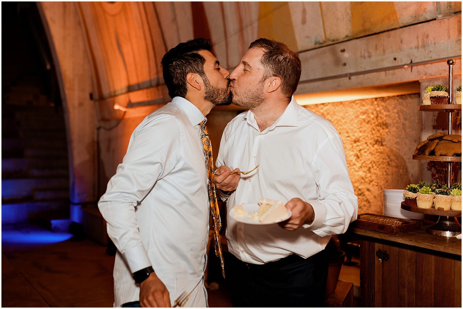 Two grooms kiss beside their cake.